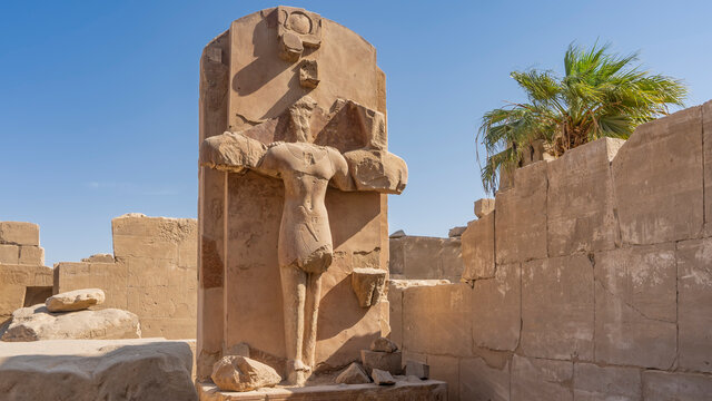 A dilapidated sculpture of Pharaoh Thutmose in the Karnak Temple of Luxor. The statue stands on a stone pedestal, against a background of blue sky. Egypt