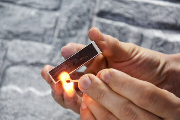 Detail of ignition of matches for a matchbox. A burning match in one hand and a matchbox in the other. The process of lighting a match. Hand with burning match on gray background