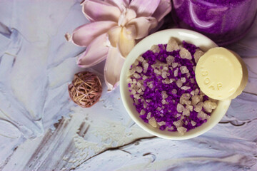 Fototapeta na wymiar Sea salt and soap. Composition of lilac sea salt in pellets, soap bar, lotus flower on white brushstrokes background flatly. Spa treatments, relaxation, feminine concept. Copy space.
