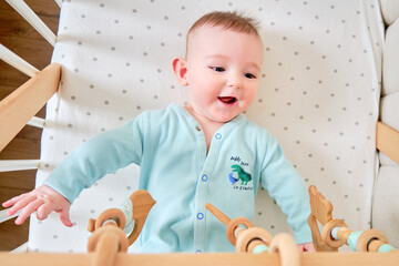Happy infant baby is playing with wooden hanging toys lying in bed. Smiling funny child in turquoise clothes in a crib, aged six months