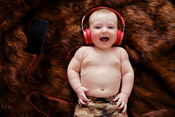 Funny happy baby boy listens to music in wired headphones online from his phone. Smiling child is lying in earphones, top view