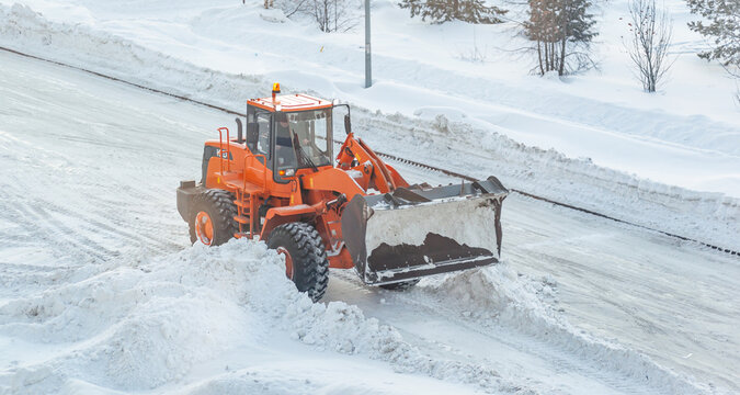 Big orange tractor cleans up snow from the road and loads it into the truck. Cleaning and cleaning of roads in the city from snow in winter. Snow removal after snowfall and blizzards. 