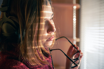 A girl with headphones behind a laptop in the sunlight, through the blinds.