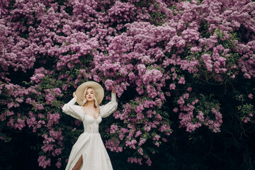 Young blonde girl in a straw hat and in a white wedding dress with an open top near the lilac bushes