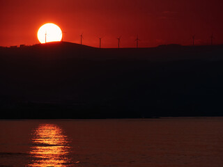 Picturesque scenery of modern windmills placed on hill on background of huge orange sun and sundown sky in evening