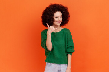 Woman with Afro hairstyle holding fingers shaped like telephone near head, communicating by phone, looking at camera with playful smile. Indoor studio shot isolated on orange background.