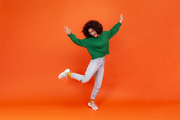 Fototapeta na wymiar Full length portrait of happy woman with Afro hairstyle wearing green casual style sweater standing on one leg, raised arms, dancing, celebrating. Indoor studio shot isolated on orange background.