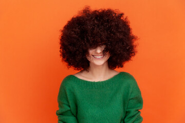 Happy positive woman with Afro hairstyle wearing green casual style sweater sowing her fluffy hair, heed hairdresser, posing with toothy smile. Indoor studio shot isolated on orange background.