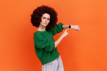 Bossy woman with Afro hairstyle wearing green casual style sweater pointing wrist watch and looking annoyed displeased, showing clock to hurry up. Indoor studio shot isolated on orange background. - obrazy, fototapety, plakaty