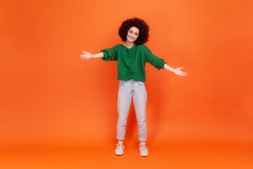 Fototapeta na wymiar Full length of woman with Afro hairstyle sharing opening hands looking at camera with kind smile, greeting and regaling, happy glad to see you. Indoor studio shot isolated on orange background.