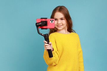 Portrait of little girl with positive expression, holding steadicam and smart phone, streaming,...