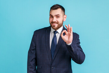 Everything is fine! Smiling bearded man wearing official style suit, looking nat camera, showing ok sign, expressing positive emotions. Indoor studio shot isolated on blue background.