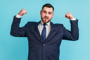 I'm strong! Portrait of self-confident motivated businessman in official style suit looking assertive and raising arms to show biceps, feeling powerful. Indoor studio shot isolated on blue background.