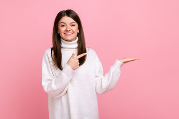 Happy woman presenting copy space on her palm, showing empty place for commercial text or goods, wearing white casual style sweater. Indoor studio shot isolated on pink background.