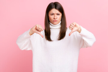 Portrait of sad brunette female showing thumb down demonstrating disapproval gesture and dislike,...