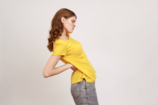 Side view portrait of exhausted unhealthy teen girl in yellow T-shirt touching aching back, suffering lower lumbar discomfort, injured spine disk. Indoor studio shot isolated on gray background.
