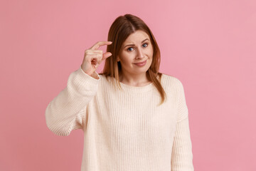 Portrait of blond woman frowning and showing a little bit gesture, asking some more, dissatisfied upset with amount, wearing white sweater. Indoor studio shot isolated on pink background.