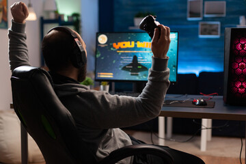 Man winning video games with joystick and headphones in front of monitor. Gamer using controller and headset, playing online games on computer. Person celebrating game win for leisure. © DC Studio