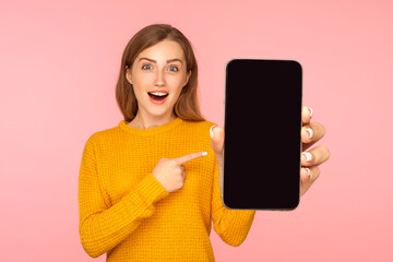 Portrait of amazed good looking girl in sweater pointing at cellphone and looking with surprise at camera, shocked about device or mobile application. indoor studio shot pink background - obrazy, fototapety, plakaty