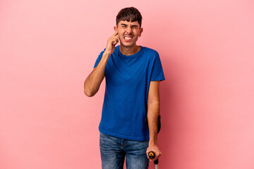 Young mixed race man with crutches isolated on pink background covering ears with hands.