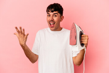 Young mixed race man holding an iron isolated on pink background receiving a pleasant surprise,...