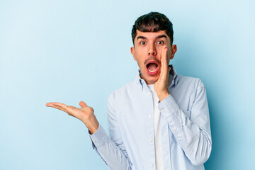 Young mixed race man isolated on blue background holds copy space on a palm, keep hand over cheek. Amazed and delighted.