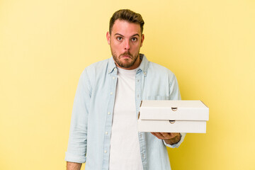 Young caucasian man holding a pizzas isolated on yellow background shrugs shoulders and open eyes confused.