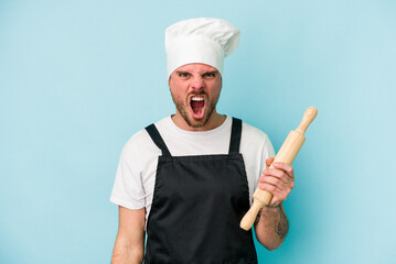 Young baker man isolated on blue background screaming very angry and aggressive.