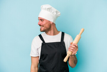 Young baker man isolated on blue background looks aside smiling, cheerful and pleasant.