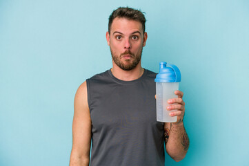 Young caucasian man drinking milkshake isolated on  blue background shrugs shoulders and open eyes confused.