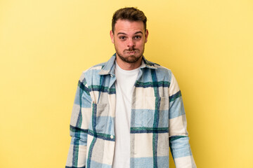 Young caucasian man isolated on yellow background blows cheeks, has tired expression. Facial expression concept.