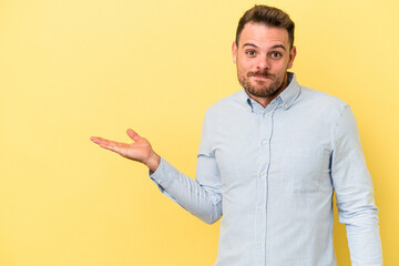 Young caucasian man isolated on yellow background showing a copy space on a palm and holding...