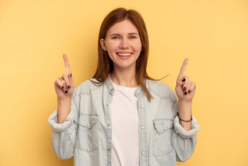 Young English woman isolated on yellow background indicates with both fore fingers up showing a blank space.