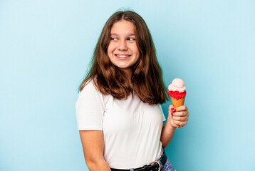 Little caucasian girl eating an ice cream isolated on blue background looks aside smiling, cheerful and pleasant.