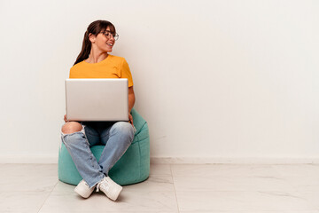 Young Argentinian woman working with pc sitting on a puff isolated on white background looks aside smiling, cheerful and pleasant.