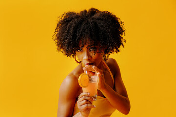 beautiful young African american woman in bright summer outfit drinking refreshing cocktail isolated on yellow background