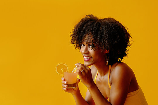 beautiful young African american woman in bright summer outfit drinking refreshing cocktail isolated on yellow background