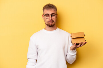 Young caucasian man holding a burger isolated on yellow background confused, feels doubtful and unsure.