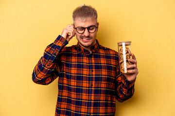 Young caucasian man holding cookies jar isolated on yellow background covering ears with hands.