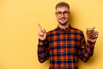 Young caucasian man holding a almond jar isolated on yellow background smiling and pointing aside, showing something at blank space.