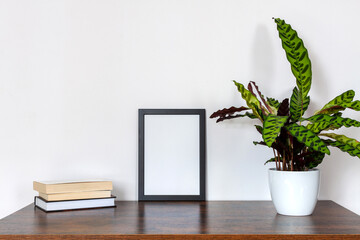 Wooden desk with houseplants. Urban jungle. White wall copy space. Black mockup frame. A stack of books. A large plant.