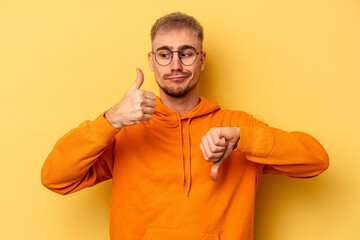 Young caucasian man isolated on yellow background showing thumbs up and thumbs down, difficult...