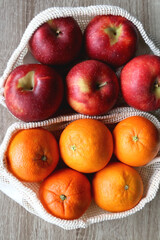 Two sustainable shopping bags filled with apples and oranges on wooden table. Top view.