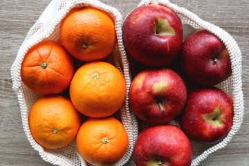 Two sustainable shopping bags filled with apples and oranges on wooden table. Top view.