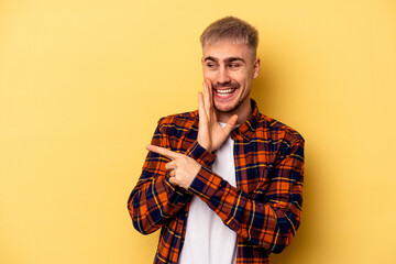 Young caucasian man isolated on yellow background saying a gossip, pointing to side reporting something.