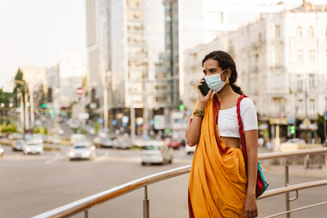 Obraz premium Young indian woman in face mask talking on cellphone at city street