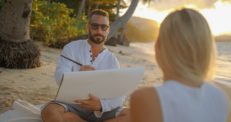 Handsome painter paints beautiful woman against sun set background of the evening ocean, honeymoon 