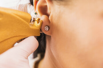 A doctor in sterile white medical gloves pierces the ears of a young girl in the medical office...