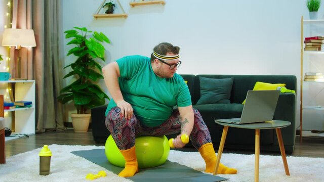 There S An Obese Man With Glasses He Has A Beard And He S Sitting On A Yellow Yoga Ball, He Is Very Concerned On Getting His Workout With Dung Bells Done, He Is Looking At The Laptop For Help