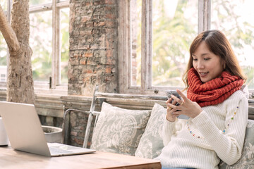 Beautiful Asian girl wearing a white sweater in winter sits on the sofa chatting online social networking apps to work from a coffee shop. 